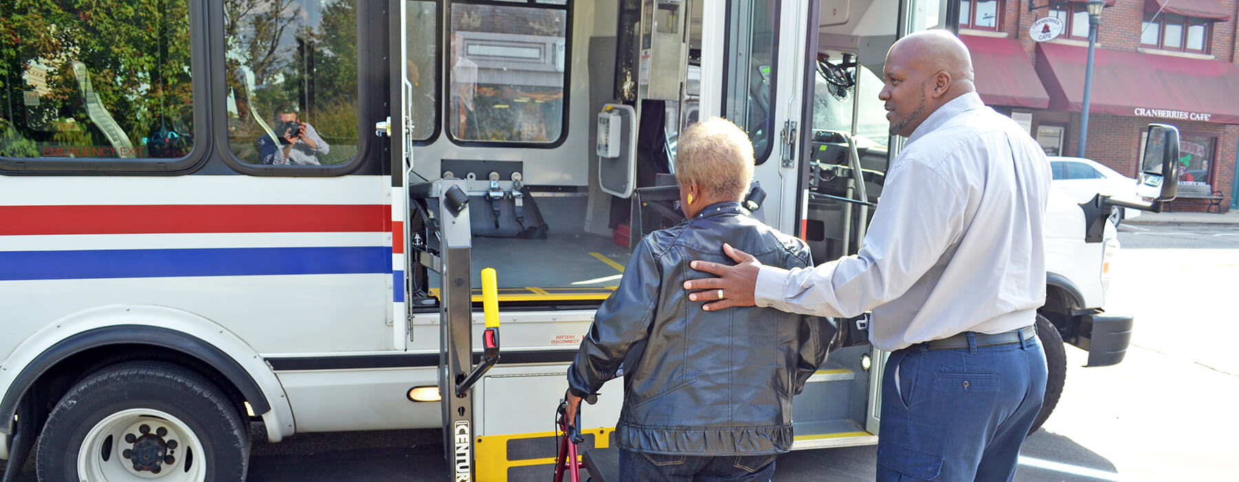 Elderly woman with walker boarding Your Ride bus with lift, assisted by the driver.