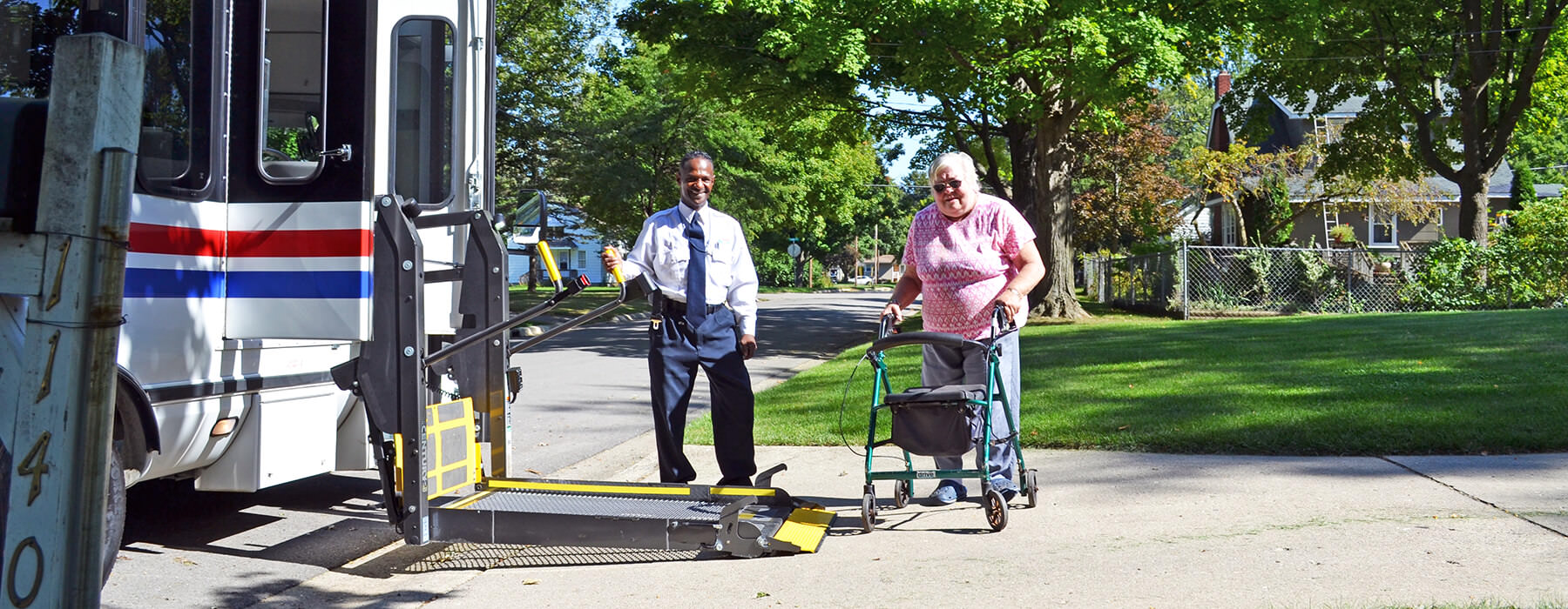 Elderly woman with walker boarding a Your Ride bus, assisted by the driver, via the wheelchair lift.